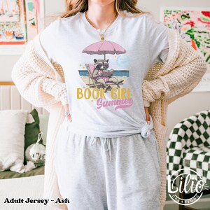 May include: A gray adult jersey t-shirt with a graphic of a raccoon wearing sunglasses and reading a book on a beach chair under an umbrella. The text "Book Girl Summer" is printed in gold on the shirt.