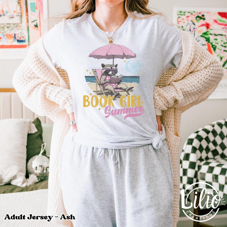 May include: A gray adult jersey t-shirt with a graphic of a raccoon wearing sunglasses and reading a book on a beach chair under an umbrella. The text "Book Girl Summer" is printed in gold on the shirt.