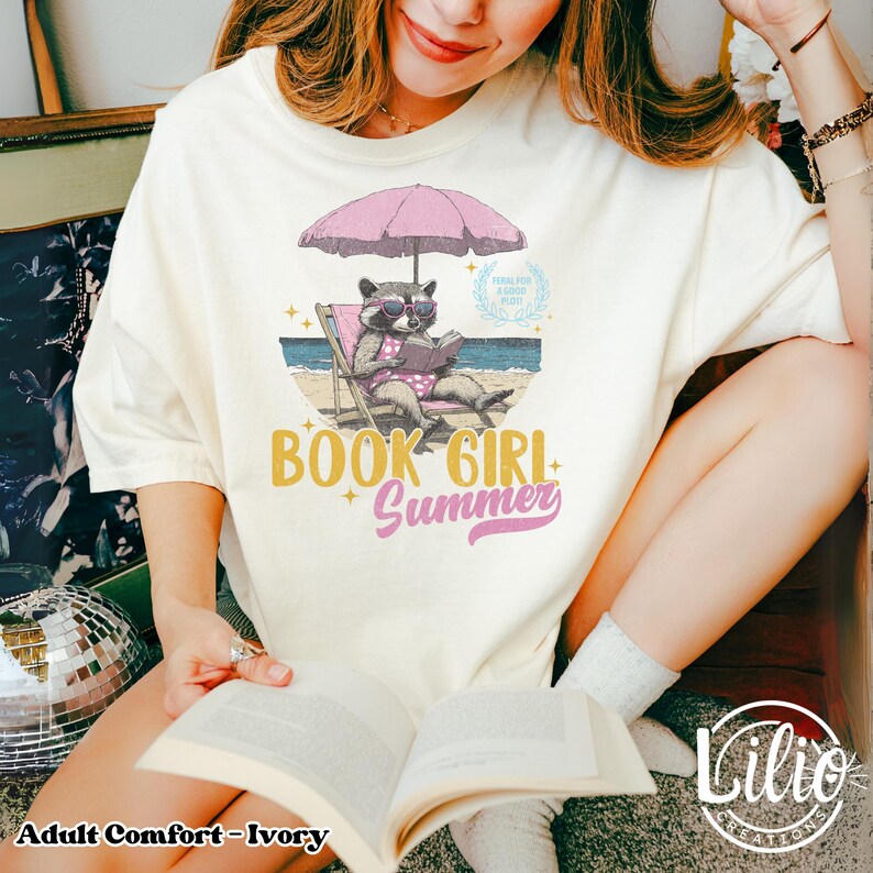 May include: A white t-shirt with a graphic of a raccoon reading a book on a beach chair under a pink umbrella. The text "Book Girl Summer" is printed on the shirt.