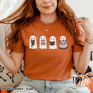 Pode incluir: Camiseta laranja com quatro fantasmas de desenho animado. Um fantasma segura um gato preto, outro livros, um terceiro l&ecirc; um livro e o &uacute;ltimo est&aacute; enrolado em um cobertor e segurando uma caneca. O texto "Adult Comfort - Yam" est&aacute; na parte inferior.