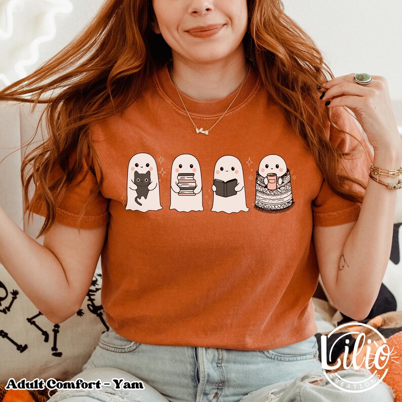 Pode incluir: Camiseta laranja com quatro fantasmas de desenho animado. Um fantasma segura um gato preto, outro livros, um terceiro l&ecirc; um livro e o &uacute;ltimo est&aacute; enrolado em um cobertor e segurando uma caneca. O texto "Adult Comfort - Yam" est&aacute; na parte inferior.