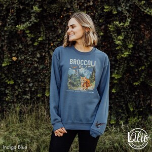 K&ouml;nnte beinhalten: Ein blaues Sweatshirt mit einem Aufdruck eines Katers mit Rucksack, der in den Bergen wandert. Der Text auf dem Sweatshirt lautet "Broccoli The Fierce" und "Official Quest Squad Mascot".