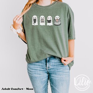 Pode incluir: Camiseta verde musgo com quatro fantasmas de desenho animado. Cada fantasma tem um design diferente: um gato preto, livros, um livro aberto e uma x&iacute;cara de caf&eacute;. O texto "Adult Comfort - Moss" est&aacute; na parte inferior esquerda.