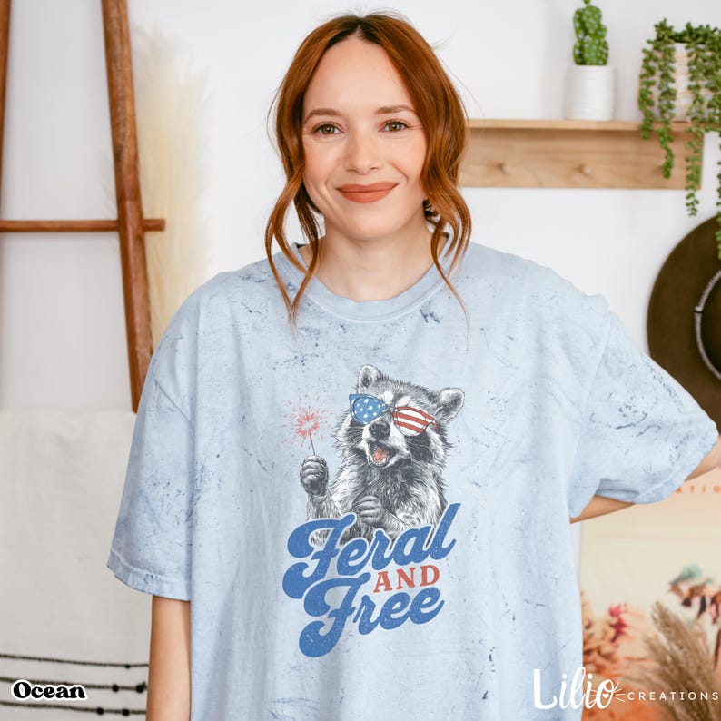 Woman wearing a light blue acid-wash T-shirt that features a playful, illustrated raccoon holding a sparkler. The raccoon wears American flag-themed sunglasses and appears mid-yell, giving off an energetic, rebellious vibe. Below the graphic, bold retro-style text reads “Feral and Free” in blue and red. The design has a patriotic Fourth of July theme and is both funny and stylish. The shirt is by Lilio Creations.