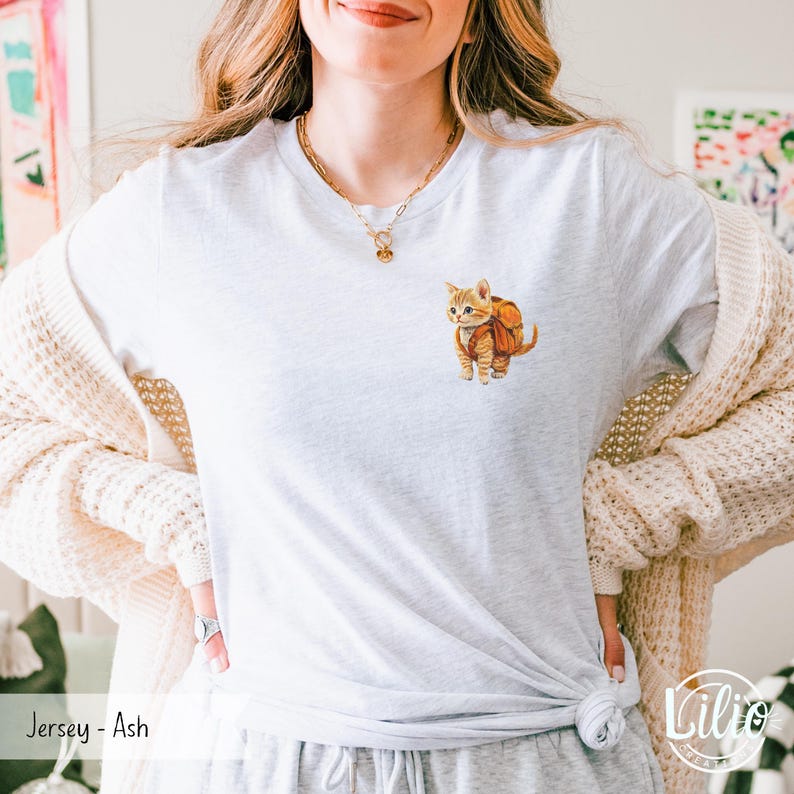 May include: A white t-shirt with a small illustration of a cat wearing a backpack. The text "Jersey - Ash" is visible at the bottom of the image.