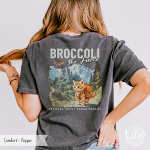 May include: A dark gray t-shirt with a vintage-style graphic of a cat wearing a backpack and the text "Broccoli The Fierce" and "Official Quest Squad Mascot".