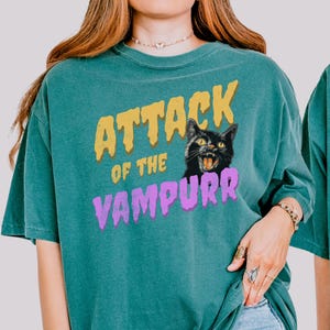 May include: A teal blue t-shirt with the text "Attack of the Vampurr" in yellow, purple, and black lettering. A black cat with yellow eyes and a menacing expression is featured in the design.