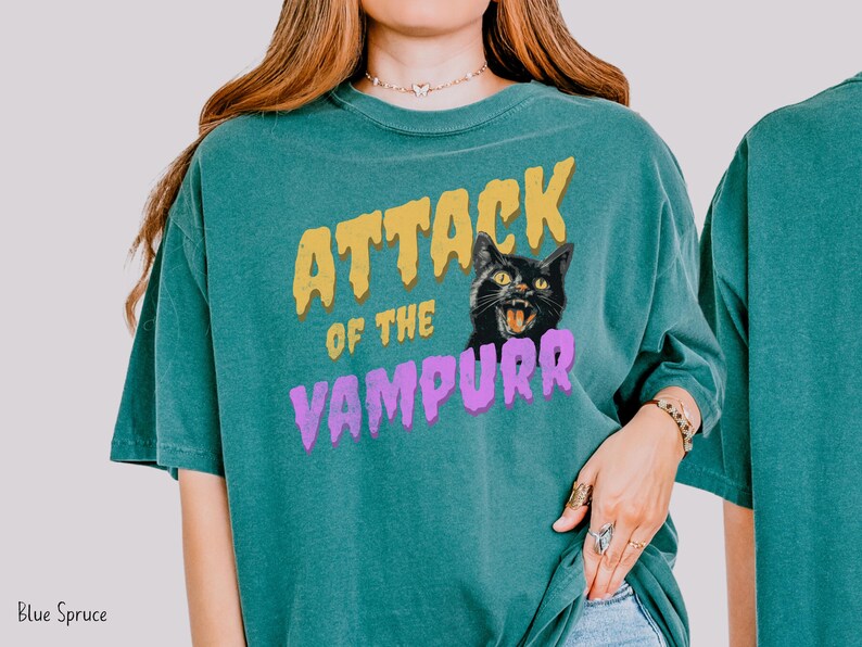 May include: A teal blue t-shirt with the text "Attack of the Vampurr" in yellow, purple, and black lettering. A black cat with yellow eyes and a menacing expression is featured in the design.