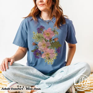 May include: Blue jean adult comfort t-shirt featuring a floral design with pink and purple flowers. The shirt has short sleeves and a crew neck. The text "Adult Comfort - Blue Jean" is at the bottom of the image.