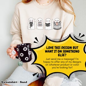Pode incluir: Camisola cor de areia com um design de quatro fantasmas de desenho animado, cada um com um acess&oacute;rio &uacute;nico: um gato preto, livros, um livro e uma caneca. A imagem inclui o texto: "LOVE THIS DESIGN BUT WANT IT ON SOMETHING ELSE?" e "Sweatshirt - Sand".