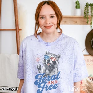 May include: A lilac t-shirt featuring a raccoon wearing sunglasses in the style of the American flag, holding a sparkler. The text "Feral and Free" is in blue script. The shirt has a relaxed fit.