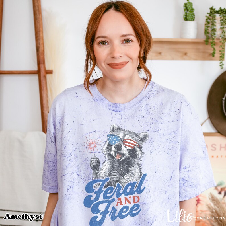 May include: A lilac t-shirt featuring a raccoon wearing sunglasses in the style of the American flag, holding a sparkler. The text "Feral and Free" is in blue script. The shirt has a relaxed fit.
