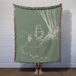May include: A green woven throw blanket with a white outline of Winnie the Pooh and Piglet walking in the woods. The blanket has a fringe border.