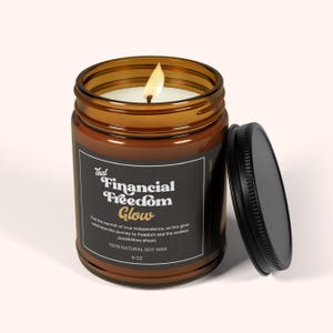 May include: A brown glass candle jar with a black label that reads "That Financial Freedom Glow". The label also includes the text "Feel the warmth of true independence, as this glow celebrates the journey to freedom and the endless possibilities ahead. 100% Natural Soy Wax 9 oz".