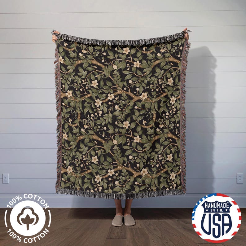 A person stands holding up a large woven blanket featuring a dark botanical design with green leaves, beige flowers, and intertwining branches on a black background. The blanket has multicolored fringe on all sides and is made from 100% cotton. It is displayed in front of a light gray shiplap wall and wood flooring. Logos in the corners note that it is “100% Cotton” and “Handmade in the USA.” Ideal for cottagecore, dark academia, and cozy home decor styles.