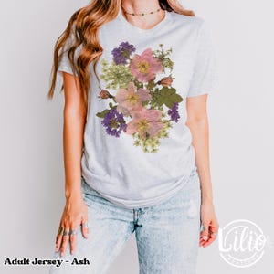 May include: Light gray adult jersey t-shirt with a floral design. The design features pink, purple, and green flowers. The text "Adult Jersey - Ash" is at the bottom left of the image.