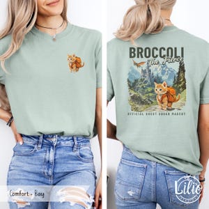 May include: A light green t-shirt with a graphic of a cat wearing a backpack and the text "Broccoli The Fierce" on the back. The cat is standing on a path in a mountainous landscape. The text "Official Quest Squad Mascot" is below the graphic.