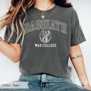 K&ouml;nnte beinhalten: Dunkelgraues T-Shirt mit dem wei&szlig;en Text "BASGIATH WAR COLLEGE". Unter dem Wort "BASGIATH" ist ein Drachenwappen zentriert. Das Shirt hat einen Rundhalsausschnitt und kurze &Auml;rmel.