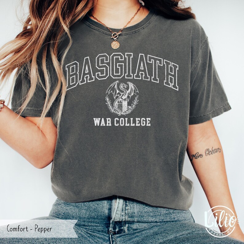 K&ouml;nnte beinhalten: Dunkelgraues T-Shirt mit dem wei&szlig;en Text "BASGIATH WAR COLLEGE". Unter dem Wort "BASGIATH" ist ein Drachenwappen zentriert. Das Shirt hat einen Rundhalsausschnitt und kurze &Auml;rmel.