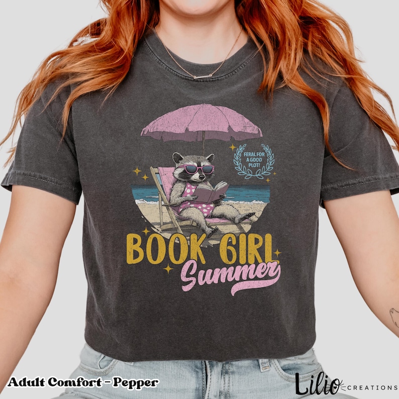 May include: A dark gray cropped t-shirt with a graphic of a raccoon wearing sunglasses and a swimsuit, reading a book under a pink umbrella on a beach. The text "BOOK GIRL SUMMER" is printed in yellow and pink on the shirt.