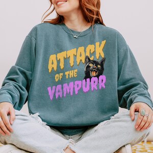 May include: A teal blue sweatshirt with the text "Attack of the Vampurr" in yellow and purple lettering. A black cat with yellow eyes and a menacing expression is featured above the text.