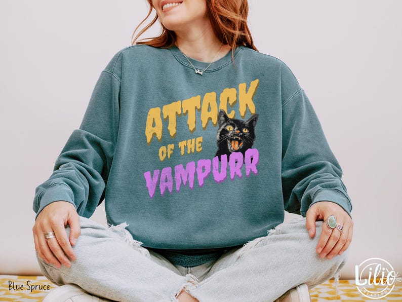 May include: A teal blue sweatshirt with the text "Attack of the Vampurr" in yellow and purple lettering. A black cat with yellow eyes and a menacing expression is featured above the text.