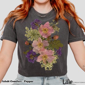 May include: Dark gray t-shirt with a floral design featuring pink and purple flowers. The text "Adult Comfort - Pepper" is at the bottom left. The shirt is a crew neck and has short sleeves. The brand name "Lilio Creations" is at the bottom right.