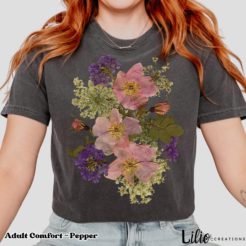 May include: Dark gray t-shirt with a floral design featuring pink and purple flowers. The text "Adult Comfort - Pepper" is at the bottom left. The shirt is a crew neck and has short sleeves. The brand name "Lilio Creations" is at the bottom right.