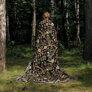 Pu&ograve; includere: Una coperta nera con un motivo floreale di fiori bianchi e foglie verdi, drappeggiata su una persona in un ambiente forestale. La coperta ha un bordo sfrangiato e un design botanico dettagliato. Lo sfondo presenta alberi ed erba verde lussureggianti.