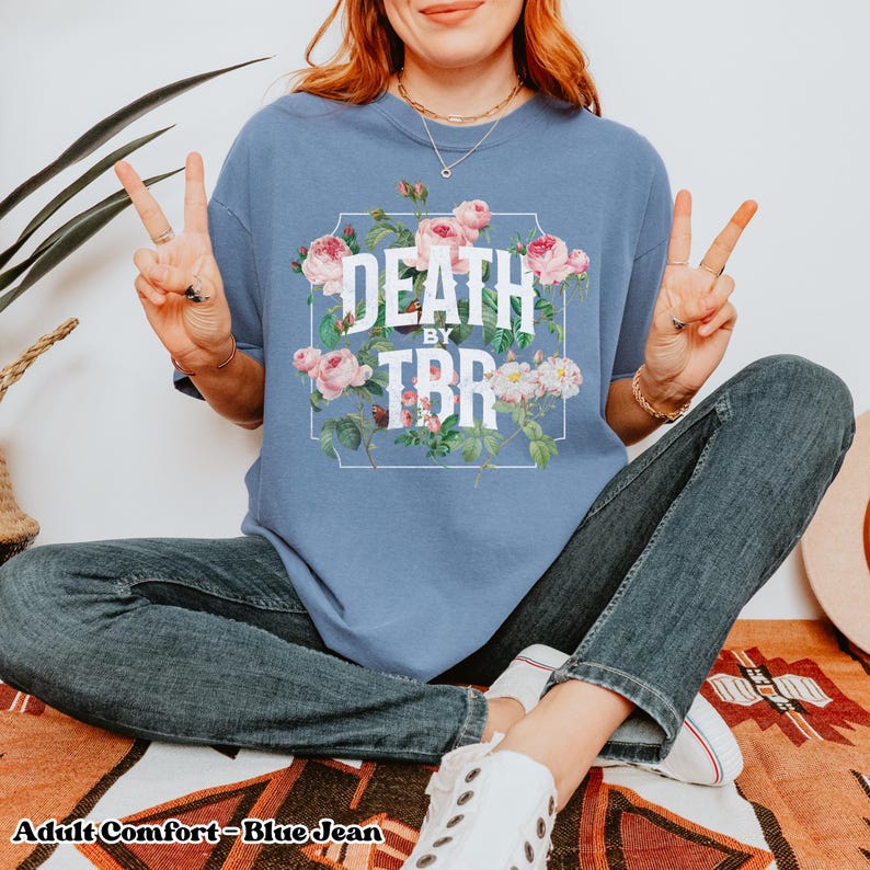 May include: A blue jean t-shirt with a floral design and the text "Death by TBR" in white. The model is wearing the shirt and sitting on a rug.