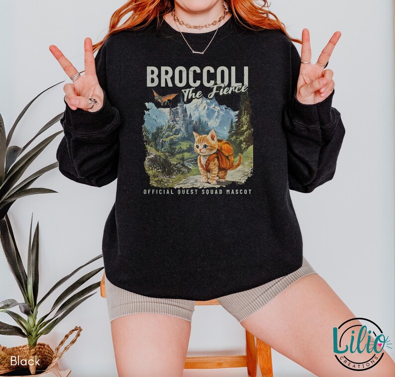 K&ouml;nnte beinhalten: Schwarzes Sweatshirt mit einem Grafikdruck eines Katers mit Rucksack und dem Text "Broccoli The Fierce" und "Official Quest Squad Mascot".