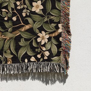 Pu&ograve; includere: Una coperta tessuta con un motivo botanico. Il design presenta fiori color crema, foglie verdi e rami marroni su sfondo nero. La coperta ha un bordo con frange multicolore.