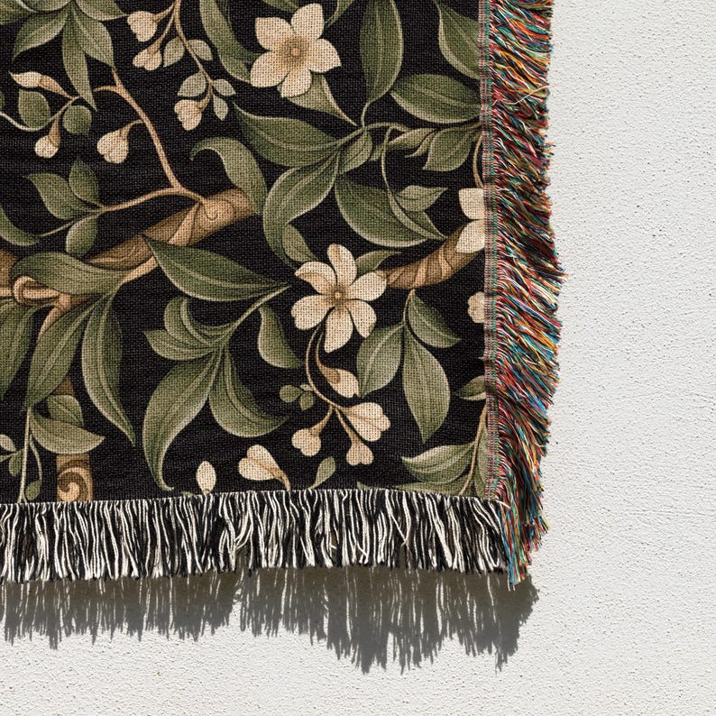 Pu&ograve; includere: Una coperta tessuta con un motivo botanico. Il design presenta fiori color crema, foglie verdi e rami marroni su sfondo nero. La coperta ha un bordo con frange multicolore.