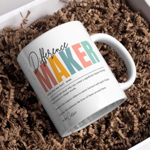 May include: White ceramic mug with the word "Difference Maker" in colorful letters and a definition of the word. The definition reads: "A dedicated individual who can create a significant impact through small actions or simple words. Someone who inspires others to move forward and spark change. A person who enhances the lives of those around them." The mug is sitting on a brown paper surface.