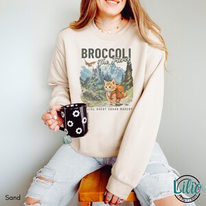 K&ouml;nnte beinhalten: Ein hellbeiger Pullover mit einem Aufdruck eines Katers mit Rucksack und dem Text "Broccoli The Fierce Official Quest Squad Mascot".