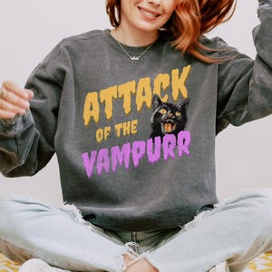 May include: A gray sweatshirt with the text "Attack of the Vampurr" in yellow and purple letters. A black cat with yellow eyes and a pink nose is featured in the design.