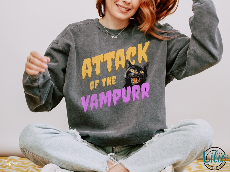 May include: A gray sweatshirt with the text "Attack of the Vampurr" in yellow and purple letters. A black cat with yellow eyes and a pink nose is featured in the design.