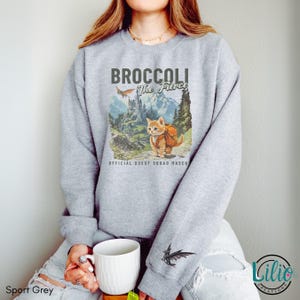 K&ouml;nnte beinhalten: Ein graues Sweatshirt mit einem Aufdruck eines Katers mit Rucksack und dem Text "Broccoli the Fierce" und "Official Quest Squad Masco".