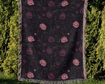 Può includere: Una coperta scura con un motivo floreale di peonie rosa e viti verdi. La coperta ha un bordo con frange. Il design presenta un motivo ripetuto di fiori grandi e piccoli. La coperta è tenuta contro una siepe verde.