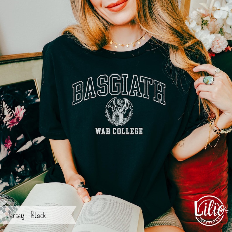 K&ouml;nnte beinhalten: Schwarzes Jersey-T-Shirt mit dem wei&szlig;en Text "BASGIATH WAR COLLEGE", der sich oben w&ouml;lbt. Ein detailliertes Emblem eines Drachen und eines Schildes befindet sich darunter. Teil einer Fantasy-Bekleidungskollektion.
