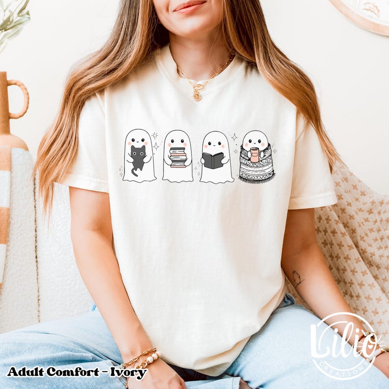 Pode incluir: Camiseta cor marfim com um design fantasioso de fantasmas. Os fantasmas s&atilde;o retratados com um gato preto, livros e uma x&iacute;cara de caf&eacute;. O texto "Adult Comfort - Ivory" &eacute; impresso na parte inferior da camiseta.