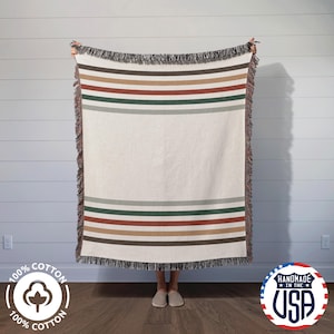 Puede incluir: Una manta tejida con un borde de flecos y un patrón de rayas en tonos marrones, verdes y beige. La manta está siendo sostenida por una persona, y el fondo es una pared blanca.