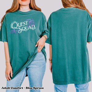 以下が含まれることがあります： 薄いターコイズブルーのTシャツに、白地に紫のアクセントで「Sounds Like a Quest Squad to Me」という文字が書かれています。ゆったりとしたフィット感で、少し大きめのシルエットです。