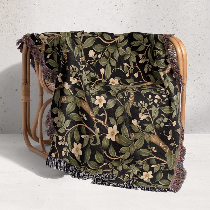 Pu&ograve; includere: Una coperta nera con un motivo botanico di foglie verdi e fiori color crema. La coperta &egrave; drappeggiata su una sedia di vimini e ha bordi sfrangiati neri, crema e rossi. Il motivo floreale &egrave; un motivo ripetuto.