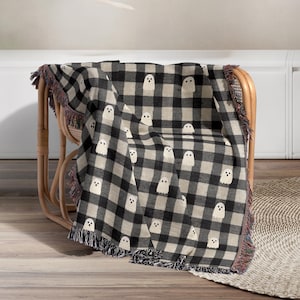 May include: A black and white checkered throw blanket with a repeating pattern of white ghost figures. The blanket has fringed edges in black, red, and tan. The blanket is draped over a wicker chair.