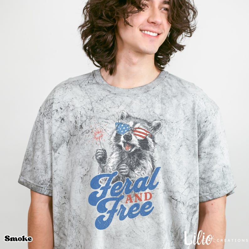 May include: A grey acid-wash t-shirt with a raccoon wearing sunglasses in the style of the American flag, holding a sparkler. The text "Feral and Free" is in blue script. The word "Smoke" is at the bottom left.