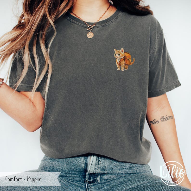 May include: A gray t-shirt with a small embroidered image of a cat wearing a backpack. The cat is orange and white. The t-shirt is a relaxed fit and has a crew neck.