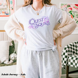 以下が含まれることがあります： 薄いグレーのTシャツに、紫と白のグラフィックデザインが施され、「Sounds like a Quest Squad to me」と書かれています。