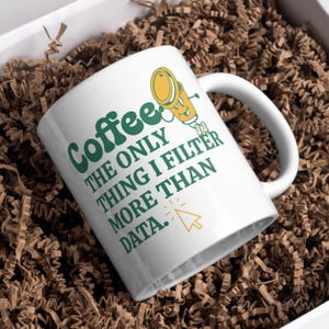 May include: White coffee mug with green text that reads "Coffee the only thing I filter more than data."  A cartoon coffee pot with a yellow body and green lid is walking to the right.  A computer mouse cursor is next to the text "data."