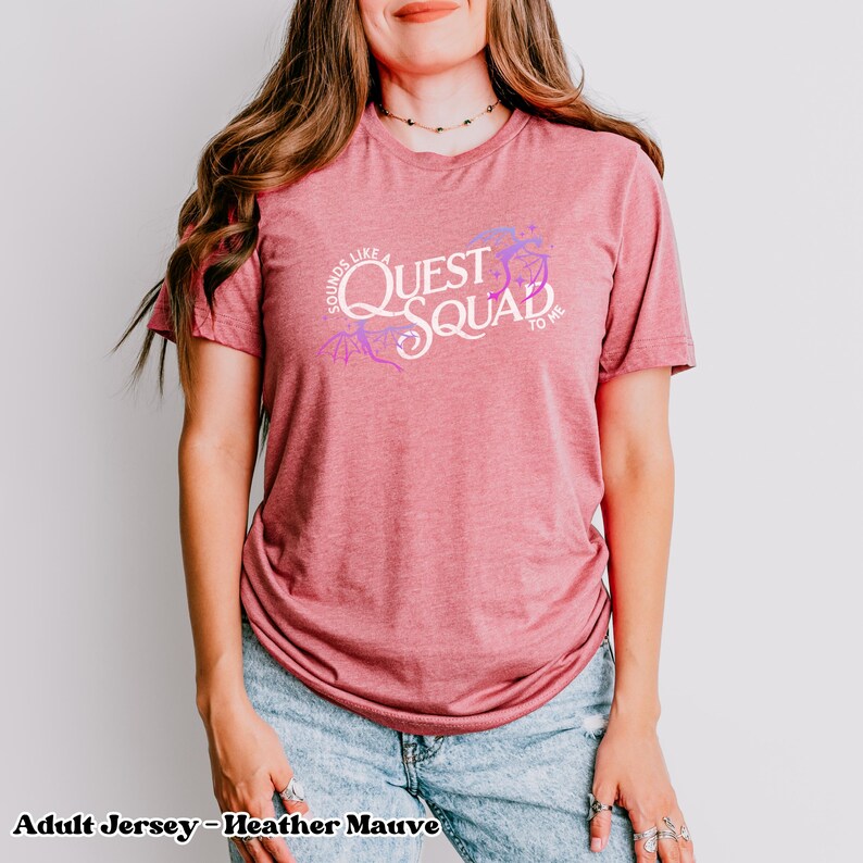 以下が含まれることがあります： ヘザーモーヴの大人用ジャージTシャツ。紫と白のグラフィックで「Sounds like a Quest Squad to me」と書いてあります。